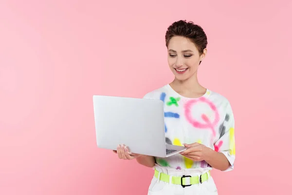 Giovane donna sorridente in abiti eleganti in piedi con computer portatile isolato su rosa — Foto stock