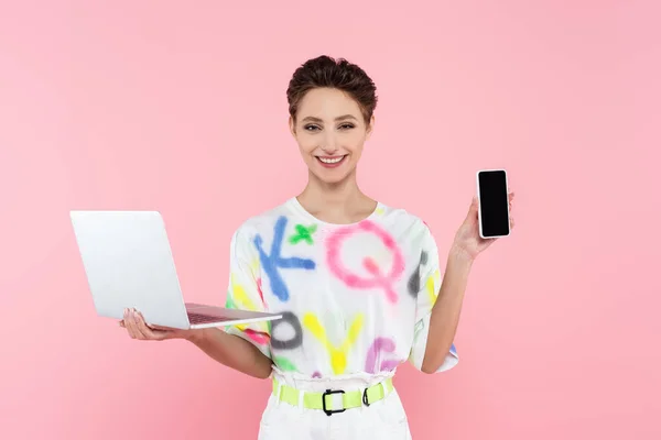 Glückliche Frau mit Laptop zeigt Handy mit leerem Bildschirm isoliert auf rosa — Stockfoto