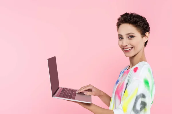 Cheerful woman looking at camera while holding laptop with blank screen isolated on pink — Stock Photo