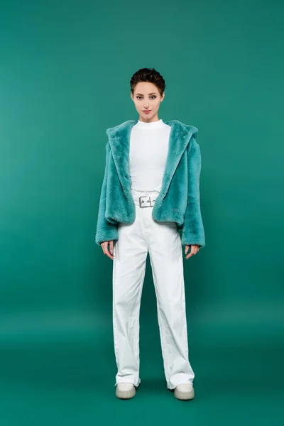 Vue pleine longueur de la femme brune en fausse fourrure veste et pantalon blanc regardant la caméra sur vert — Photo de stock