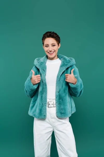 Mujer alegre en la chaqueta de piel sintética de moda que muestra los pulgares hacia arriba aislado en verde - foto de stock