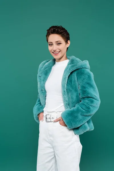 Donna soddisfatta in giacca di eco pelliccia in piedi con le mani in tasche di pantaloni bianchi isolati su verde — Foto stock