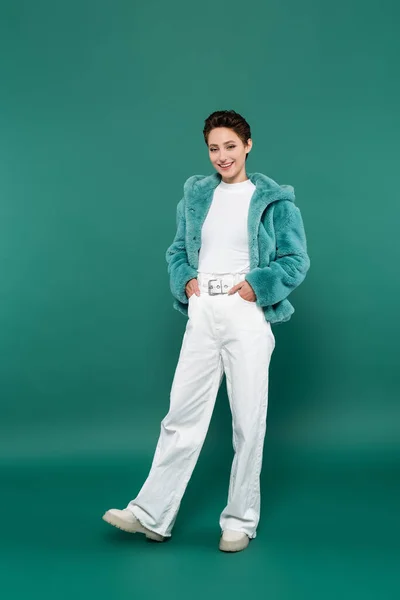 Happy and stylish woman in faux fur jacket posing with hands in pockets of white trousers on green — Stock Photo