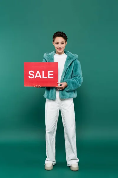 Vista a figura intera di donna felice in pantaloni bianchi e giacca di pelliccia sintetica con cartellino rosso con scritte in vendita sul verde — Foto stock