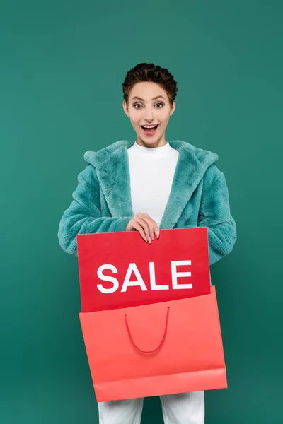 Astonished woman holding red sale card and pink shopping bag isolated on green — стоковое фото