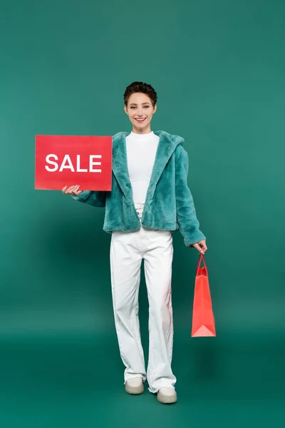 Mujer feliz en chaqueta de piel sintética y pantalones blancos con bolsa de compras y tarjeta roja con letras de venta en verde - foto de stock