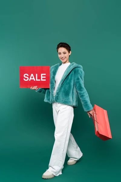 Cheerful woman in white trousers and faux fur jacket walking with sale card and shopping bag on green — Stock Photo