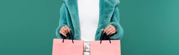 Cropped view of woman in trendy faux fur jacket holding pink shopping bags isolated on green, banner — стоковое фото