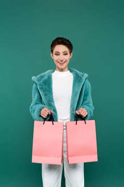 Brunette woman in faux fur jacket smiling at camera while holding pink shopping bags isolated on green — Stockfoto