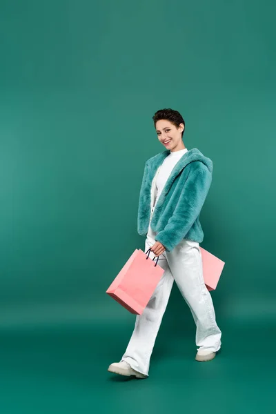 Joyful woman in faux fur jacket and white trousers walking with pink shopping bags on green — стоковое фото