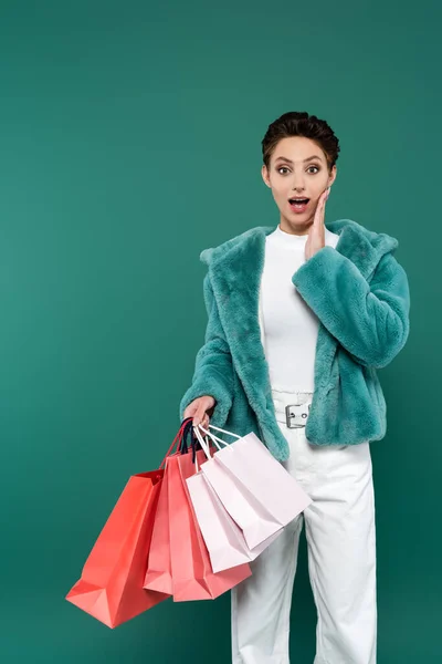 Здивована жінка в модному одязі, торкаючись обличчя, тримаючи різнокольорові сумки для покупок ізольовані на зеленому — стокове фото
