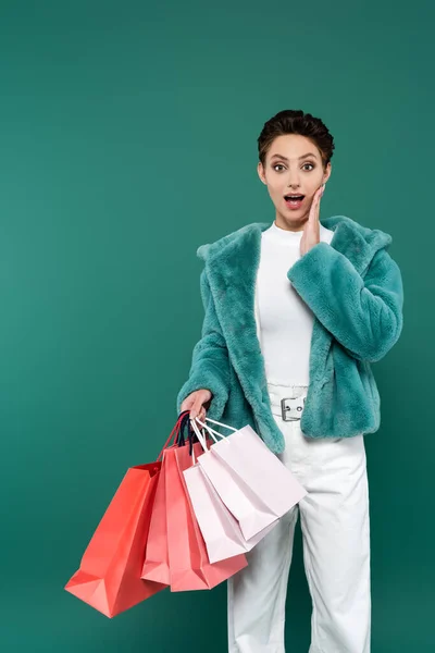 Femme étonnée dans des vêtements à la mode touchant le visage tout en tenant des sacs à provisions multicolores isolés sur vert — Photo de stock