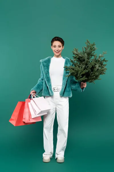 Full length view of trendy woman with shopping bags and small fir tree smiling at camera on green — стоковое фото