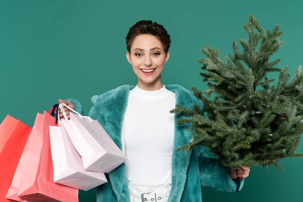 Happy and stylish woman with shopping bags and small fir tree looking at camera isolated on green — Foto stock