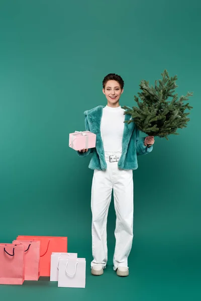 Повний вигляд задоволеної жінки з подарунковою коробкою та маленьким сосновим деревом біля сумки для покупок на зеленому — стокове фото