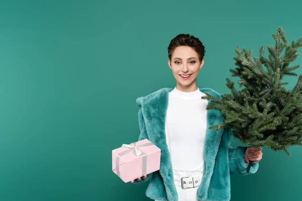 Mulher da moda com pequena árvore de abeto e caixa de presente sorrindo para a câmera isolada no verde — Fotografia de Stock