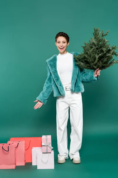 Full length view of happy and trendy woman with small christmas tree pointing at shopping bags on green — Fotografia de Stock