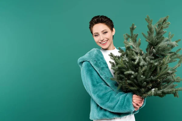 Mujer alegre en chaqueta de piel sintética de moda sosteniendo pequeño árbol de Navidad aislado en verde - foto de stock
