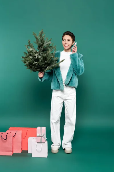 Mujer de moda y feliz con abeto pequeño hablando en el teléfono móvil cerca de bolsas de compras en verde - foto de stock