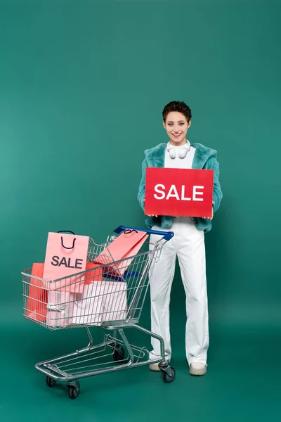 Volle Länge Ansicht der glücklichen Frau mit Verkaufskarte in der Nähe Einkaufswagen mit Einkäufen auf grün — Stockfoto