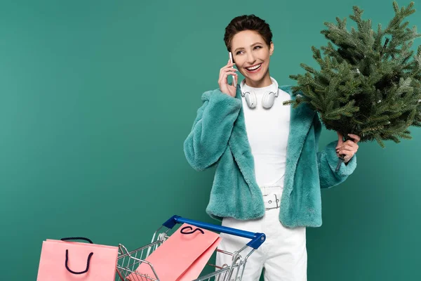 Donna sorridente con piccolo abete che parla su smartphone vicino al carrello con borse della spesa isolate sul verde — Foto stock