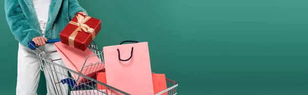 Partial view of trendy woman with present near shopping cart with purchases isolated on green, banner — Stock Photo