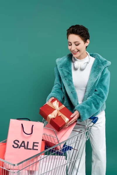 Brunette woman in faux fur jacket holding gift box near shopping cart with purchases isolated on green — Foto stock
