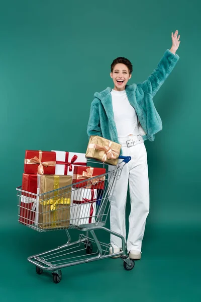 Full length view of excited woman in trendy clothes waving hand near shopping trolley with gift boxes on green — Stockfoto