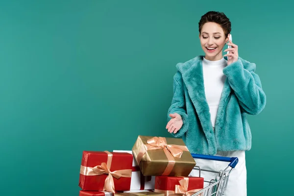 Stylish woman smiling while talking on smartphone and pointing at gift boxes in shopping trolley isolated on green — Foto stock