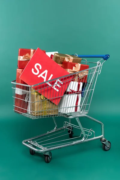 Card with sale lettering and gift boxes in shopping trolley on grey background — Stock Photo