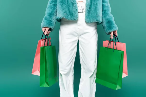 Vista parcial de la mujer en chaqueta de piel sintética y pantalones blancos sosteniendo bolsas de compras multicolores aislados en verde - foto de stock