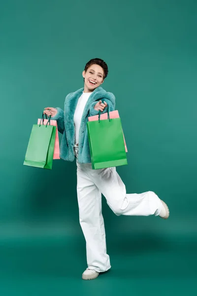 Full length view of excited woman in faux fur jacket and white trousers posing with shopping bags on green — стоковое фото