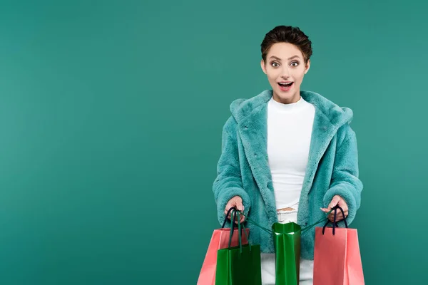 Erstaunt brünette Frau in Kunstpelzjacke mit Einkaufstaschen, während sie isoliert auf grün in die Kamera schaut — Stockfoto