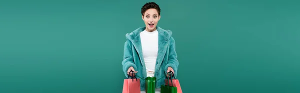 Astonished woman in faux fur jacket looking at camera while holding shopping bags isolated on green, banner — Fotografia de Stock