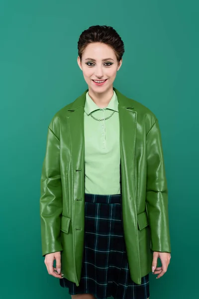 Щаслива брюнетка в шкіряній куртці і плетеній спідниці дивиться на камеру ізольовано на зеленому — стокове фото