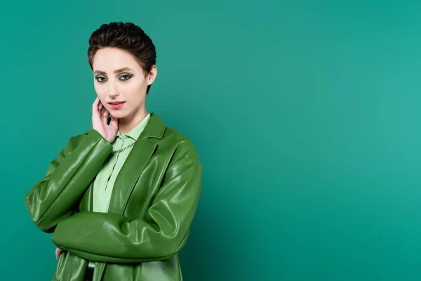 Mujer bonita en ropa de moda sosteniendo la mano cerca de la cara mientras mira a la cámara en verde - foto de stock