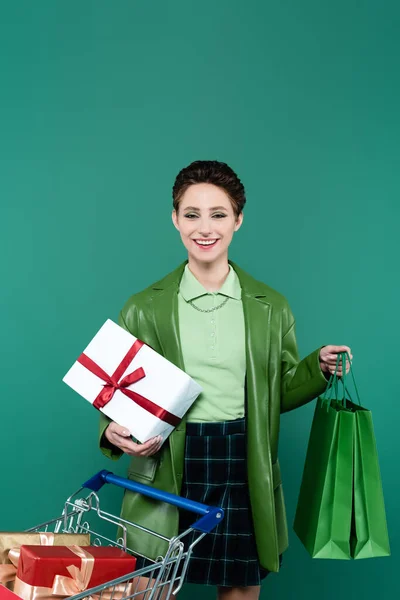 Cheerful and trendy woman with purchases near shopping cart with gift boxes isolated on green — Foto stock
