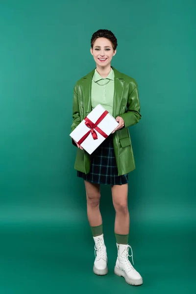 Full length view of happy woman in leather jacket, checkered skirt and white boots posing with gift box on green — Stockfoto