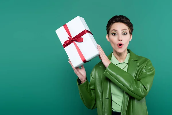 Astonished woman in trendy jacket holding white gift box and looking at camera on green — Foto stock