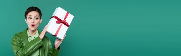 Amazed brunette woman looking at camera while holding white gift box isolated on green, banner — Stock Photo