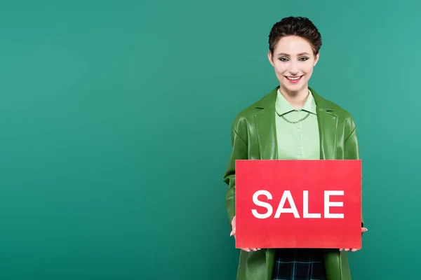 Mujer complacida en chaqueta de cuero con tarjeta roja con letras venta aislado en verde - foto de stock
