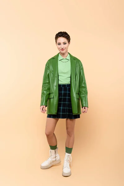 Full length view of positive woman in green jacket, plaid skirt and white lace-up boots on beige — Stock Photo