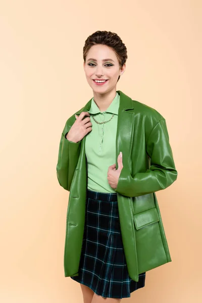 Brunette woman in green leather jacket and plaid skirt smiling at camera isolated on beige — Stockfoto
