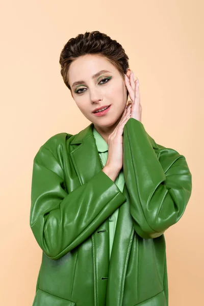 Красивая брюнетка в модном зеленом пиджаке позирует с руками рядом с лицом, изолированным от бежевого — стоковое фото