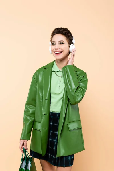 Joyful and fashionable woman with shopping bags adjusting headphones isolated on beige — Stockfoto