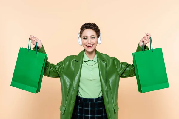 Mujer excitada en auriculares y chaqueta verde que sostiene compras aisladas en beige - foto de stock