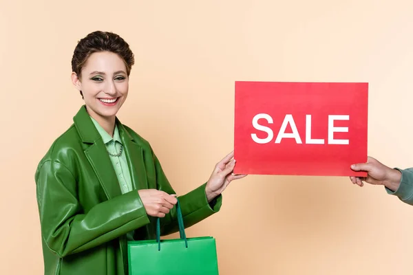 Mujer de moda con bolsas de compras cerca del hombre que sostiene la tarjeta con letras de la venta en beige - foto de stock