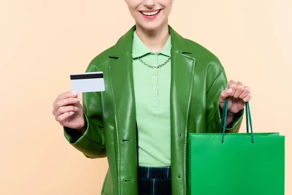 Vue recadrée de femme gaie en veste en cuir vert tenant carte de crédit et sac à provisions isolé sur beige — Photo de stock