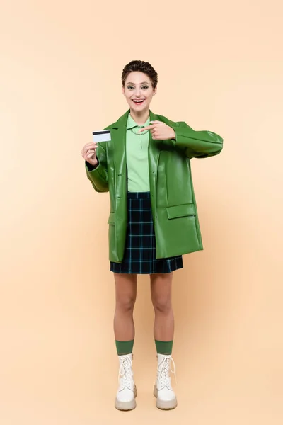 Vista completa de la mujer alegre en falda a cuadros y chaqueta verde apuntando a la tarjeta de crédito en beige - foto de stock