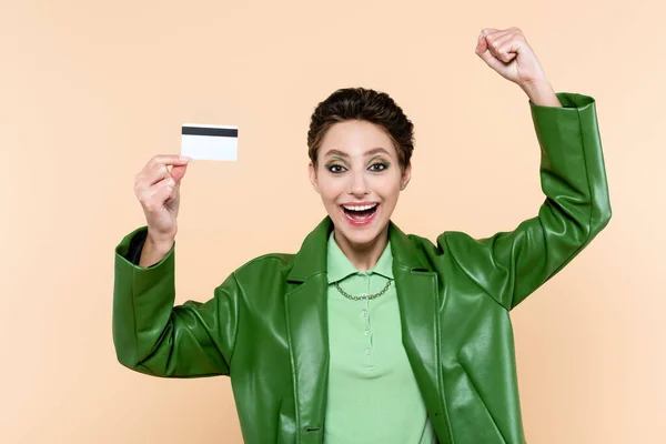 Excited woman in green leather jacket showing success gesture while holding credit card isolated on beige — Fotografia de Stock